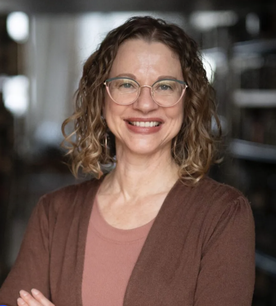 Smiling woman with curly shoulder-length hair and glasses, wearing a brown cardigan over a rose-colored top, standing with arms crossed in a softly lit indoor setting.